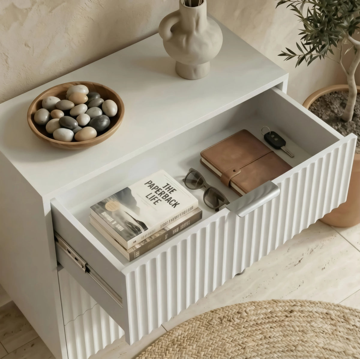 [White] flutrina design nightstand displaying an open drawer with organised books, glasses inside, and decor items on top