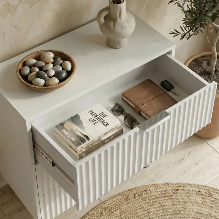 [White] flutrina design nightstand displaying an open drawer with organised books, glasses inside, and decor items on top