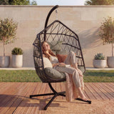 [Black] Woman seated in black hanging egg chair outdoors with pale stone wall and tall planters of green shrubs behind