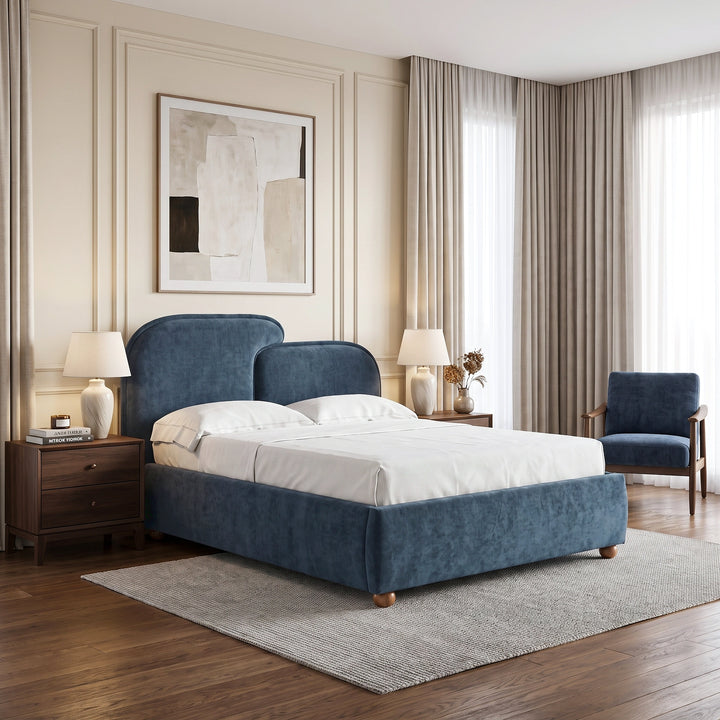 [Double | Blue] upholstered bed with a rounded headboard and footboard stands in a styled bedroom. The bed is dressed in white bedding, placed on a light gray rug between two wooden nightstands with lit table lamps.