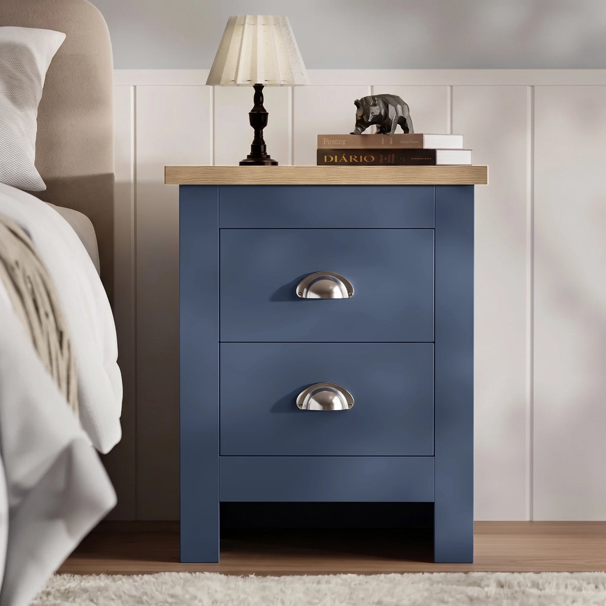 [Blue] Bedside table front view beside a bed, with lamp, stacked books, oak top, panelled wall, and rug