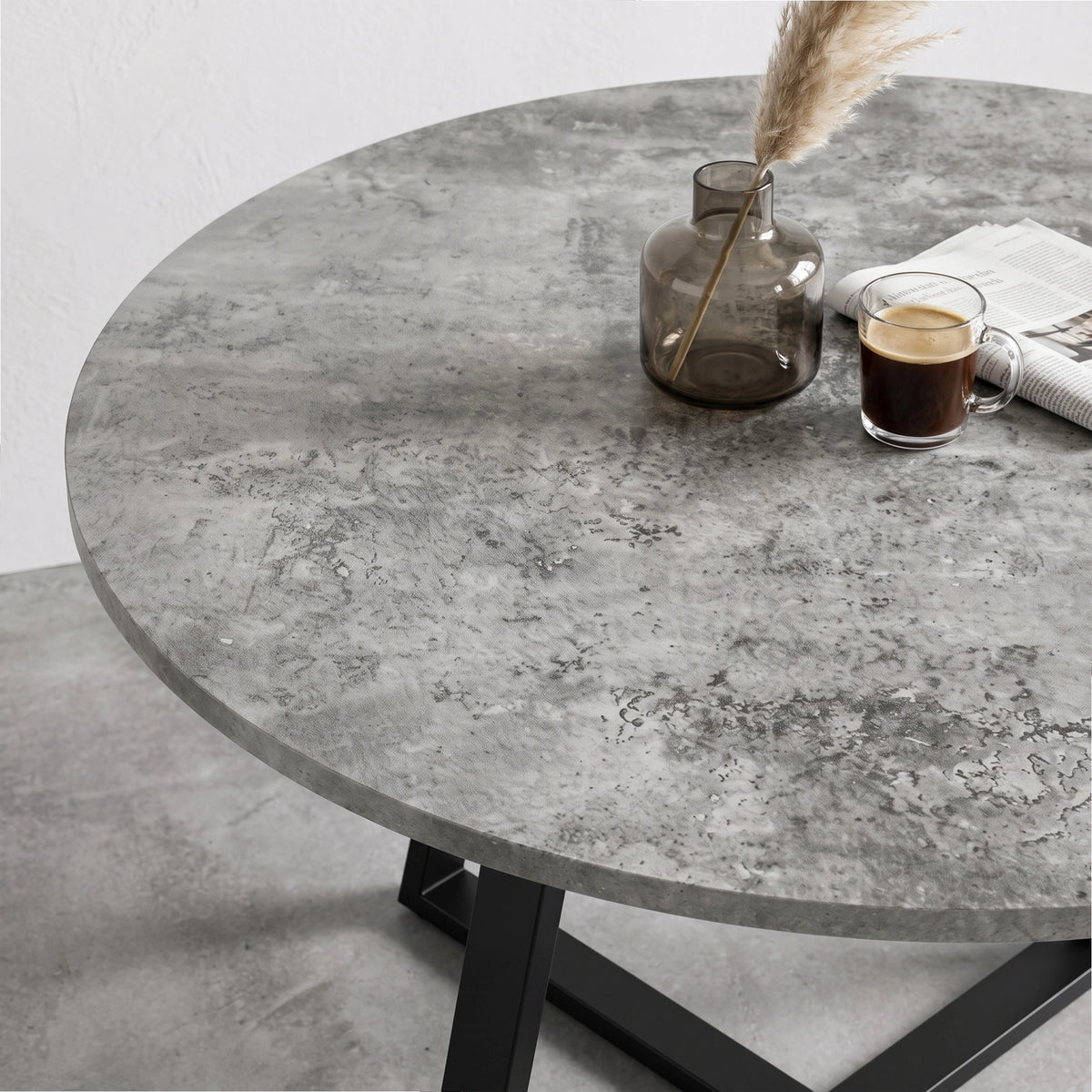 [Concrete] Close-up of the round concrete-effect tabletop with mottled patterning, styled with a vase, coffee and paper.