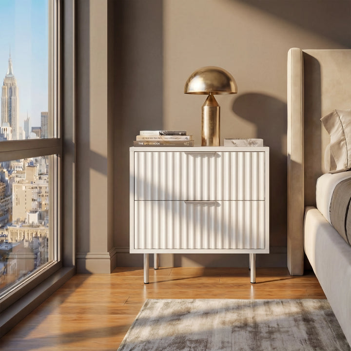 [White] flutrina two drawer bedside table with metal handles placed beside a bed in a softly lit bedroom