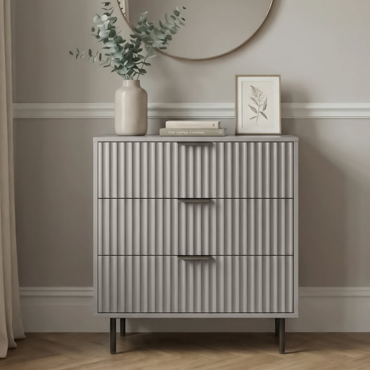 [Grey] chest of drawers with three storage drawers and flutrina front detailing placed in a bedroom