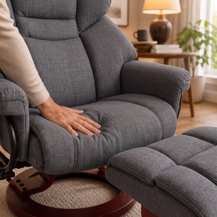 [Grey - Linen] upholstered recliner chair with a matching ottoman in a warm living room. A person's hand rests on the chair's seat cushion, showing comfort and texture.