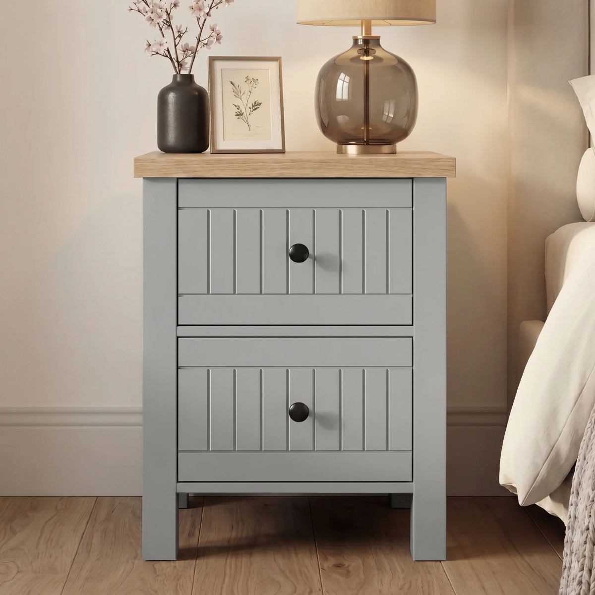 [Grey] Linra Painted 4 Piece Bedroom Furniture Set Grey bedside table with two drawers styled beside a bed with a smoked glass lamp, botanical print, and blossom stem vase