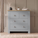 [Grey] Four drawer chest front view with oak top, chrome cup handles, pampas vase, books, and panelled wall