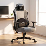 [Grey] ergonomic gaming chair with matching headrest and lumbar pillows, shown in a bright home office setting with a desk, computer monitor, and window.