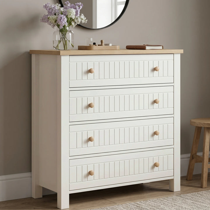 [White] Linra 4 drawer chest angled view with oak top styled with dried floral vase, sculptural ornament, and stacked books below round mirror
