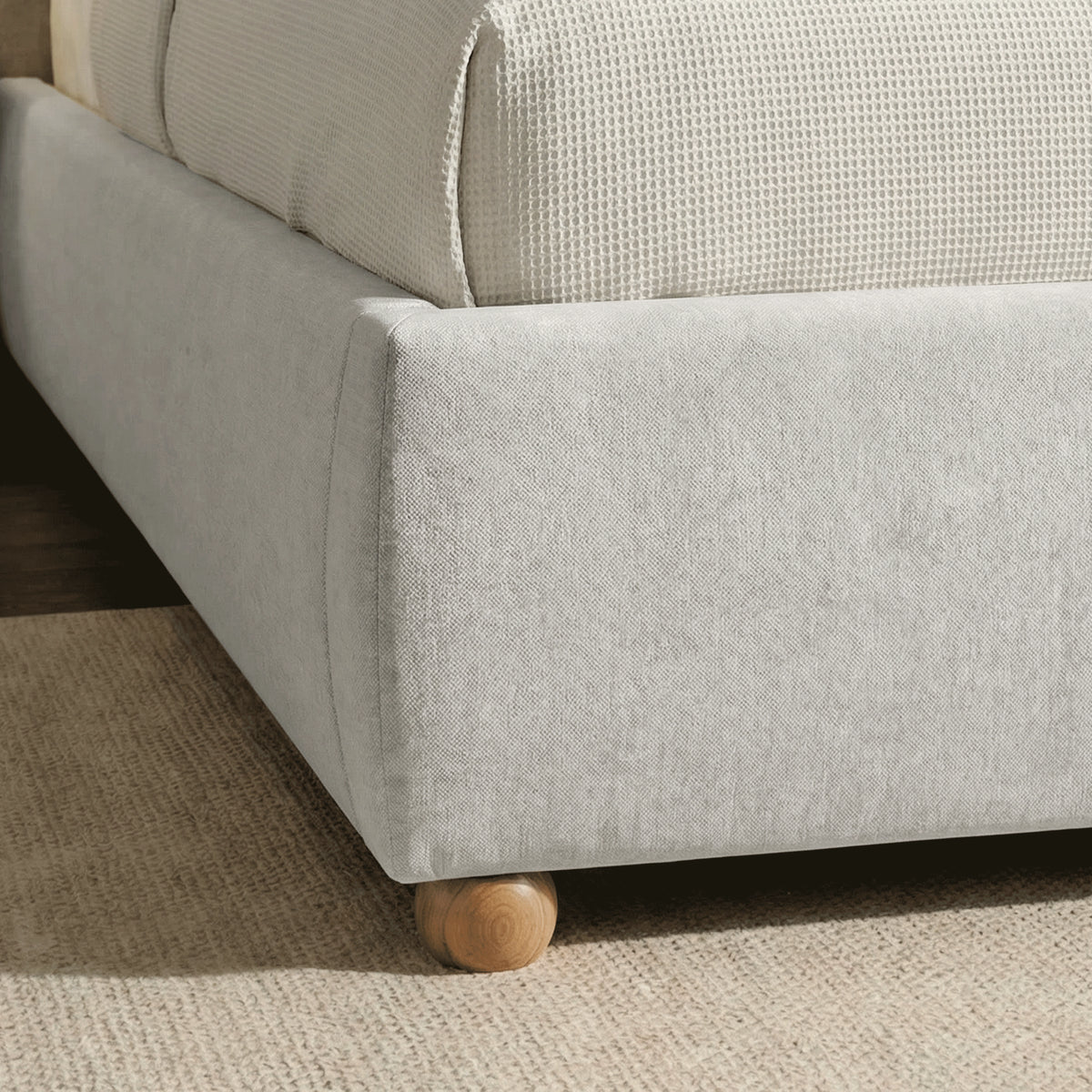 [Grey | King] Bed frame foot detail showing single round wooden leg and padded base with textured throw visible above