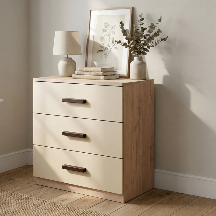 [Oak + Cashmere] Wooden dresser with three drawers, a lamp, books, and a plant in a room with a neutral color scheme.