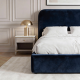 [King | Blue] King Pluva ottoman bed blue side perspective showing curved headboard silhouette beside lit lamp