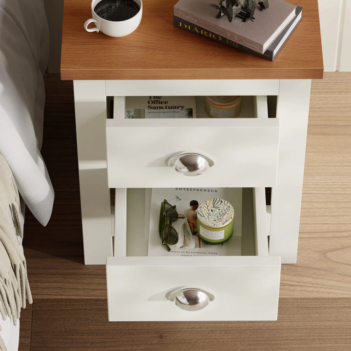 [White] Open drawers of white 2 drawer bedside table revealing storage space with candles and books inside, placed next to a bed.