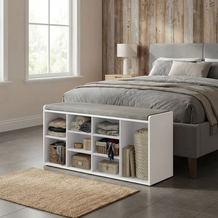 [White] Shoe storage bench at foot of grey upholstered bed with woven baskets and folded linens in cubbies