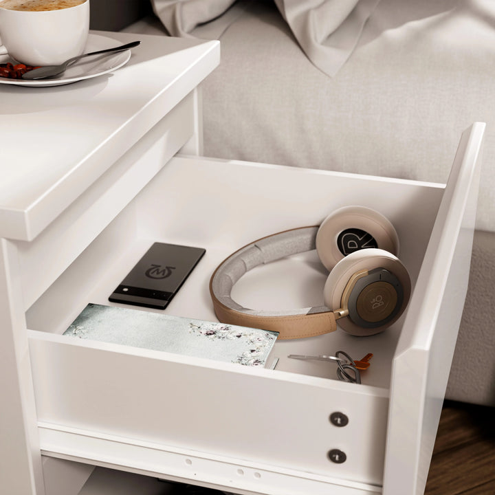 [White] close-up of open drawer in white bedside table showing phone headphones notebook and keys