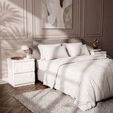 [White] A neatly made bed with white bedding and pillows in a styled bedroom, with soft sunlight casting shadows across the room.