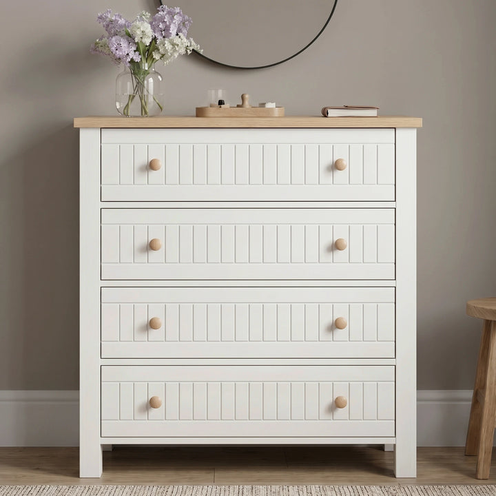[White] Linra 4 drawer chest front view with oak top styled with dried floral vase, sculptural ornament, and books below a round black mirror