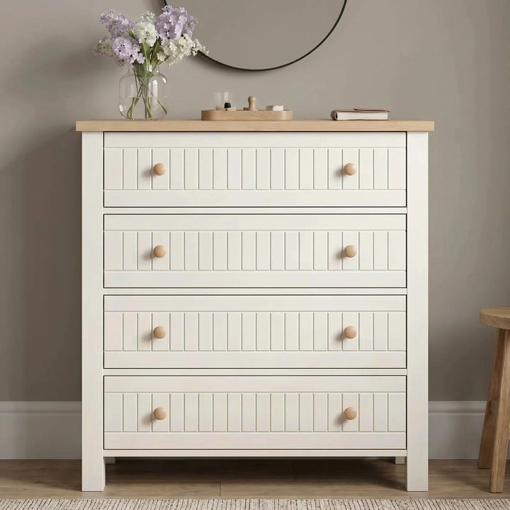 [White] Linra White Painted 4 Piece Bedroom Set four-drawer chest front view with tongue-and-groove fronts, paired round wooden knobs, oak top, and lilac flowers beside a round mirror