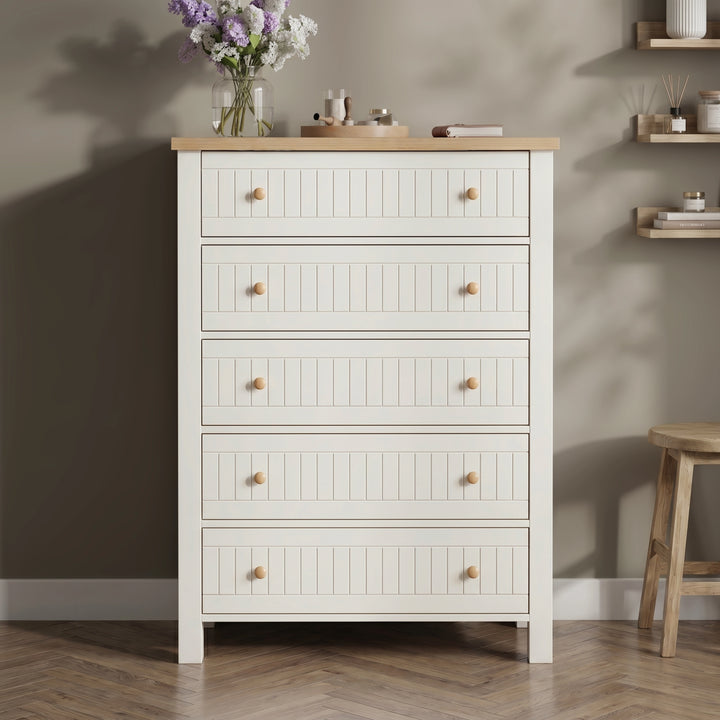 [White] Linra tall 5 drawer chest front view against dark wall with oak top styled with pale vase, dried palm, and brass candle tray