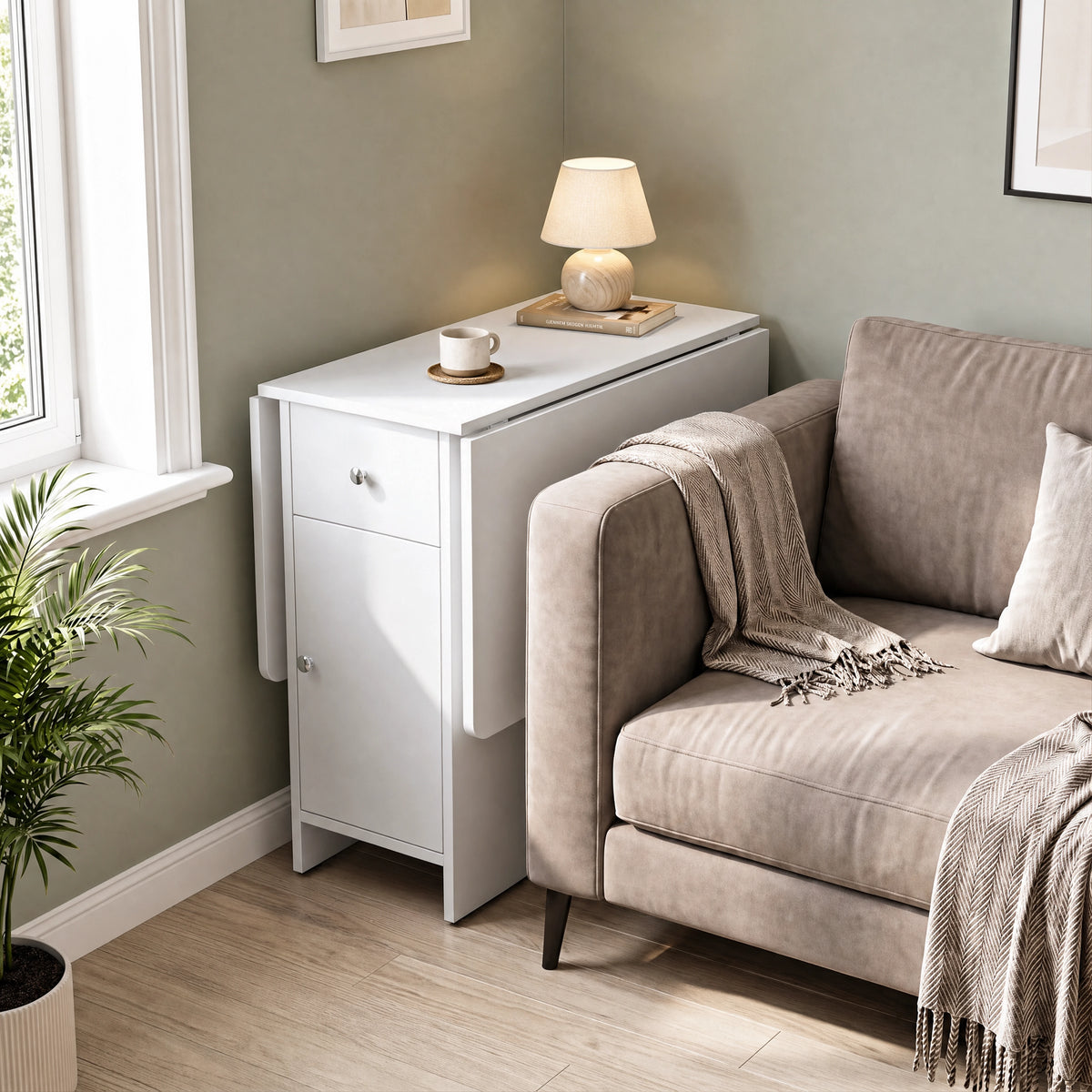 [White] Small folding dining table closed beside a sofa and window, featuring white cabinet & top with lamp, books and a mug.